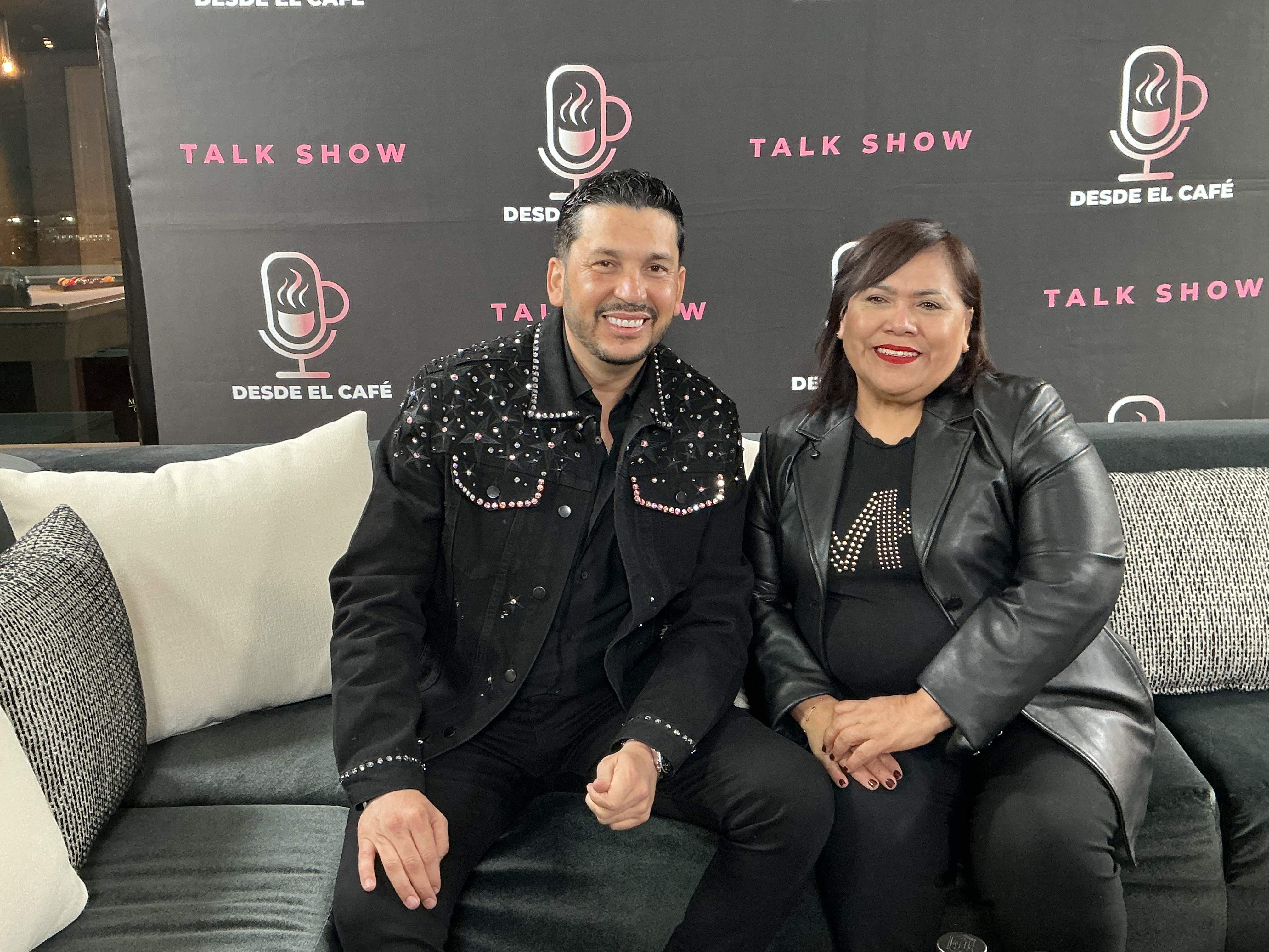 Conductora y artista invitado sentados frente al muro de prensa del show